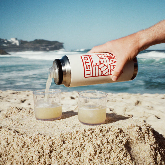 A hand pours a pale yellow drink from a beige Lister insulated bottle with red logo graphics into two clear cups on a sandy beach with the ocean and blue sky in the background.