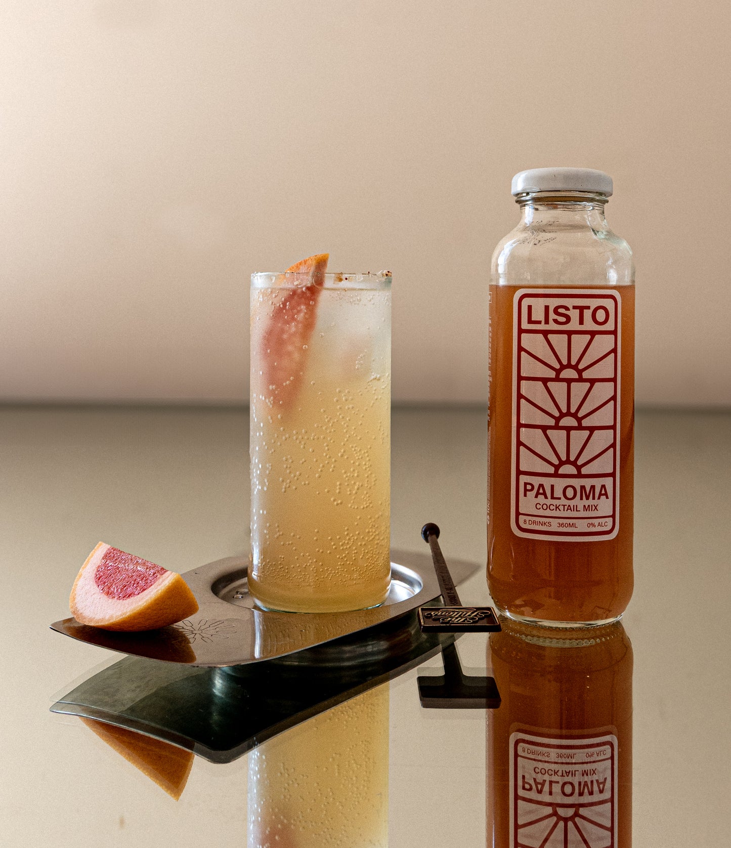 Cocktail with a slice of grapefruit next to a bottle labeled 'Listo Paloma' on a reflective surface.