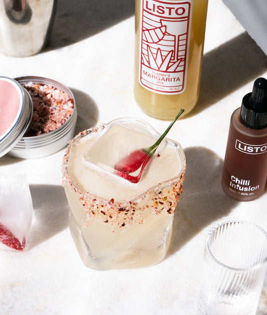 Margarita with a chilli garnish, Listo margarita bottle, and Listo chili infusion bottle on a white surface.